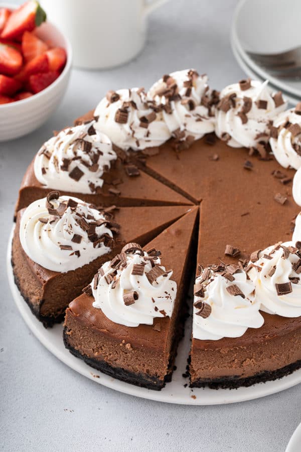 Sliced chocolate cheesecake topped with whipped cream on a white cake plate