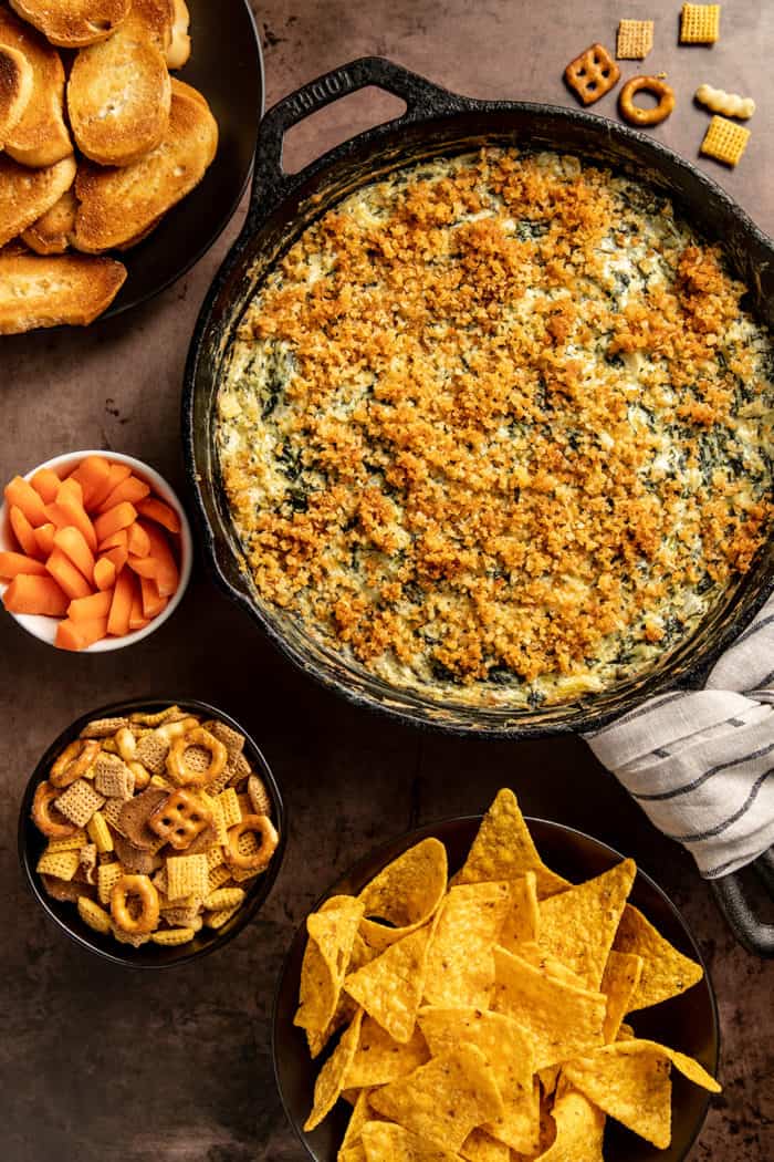 Freshly baked spinach artichoke dip in a skillet surrounded by chips, bread, and carrots