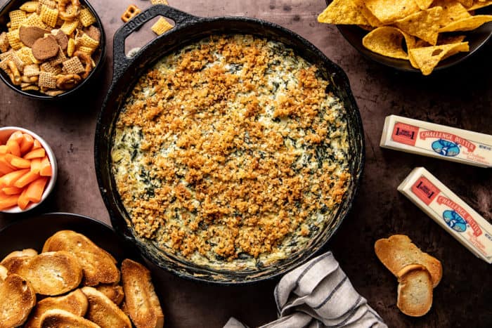 Skillet with spinach artichoke dip on a counter surrounded by a variety of chips and bread