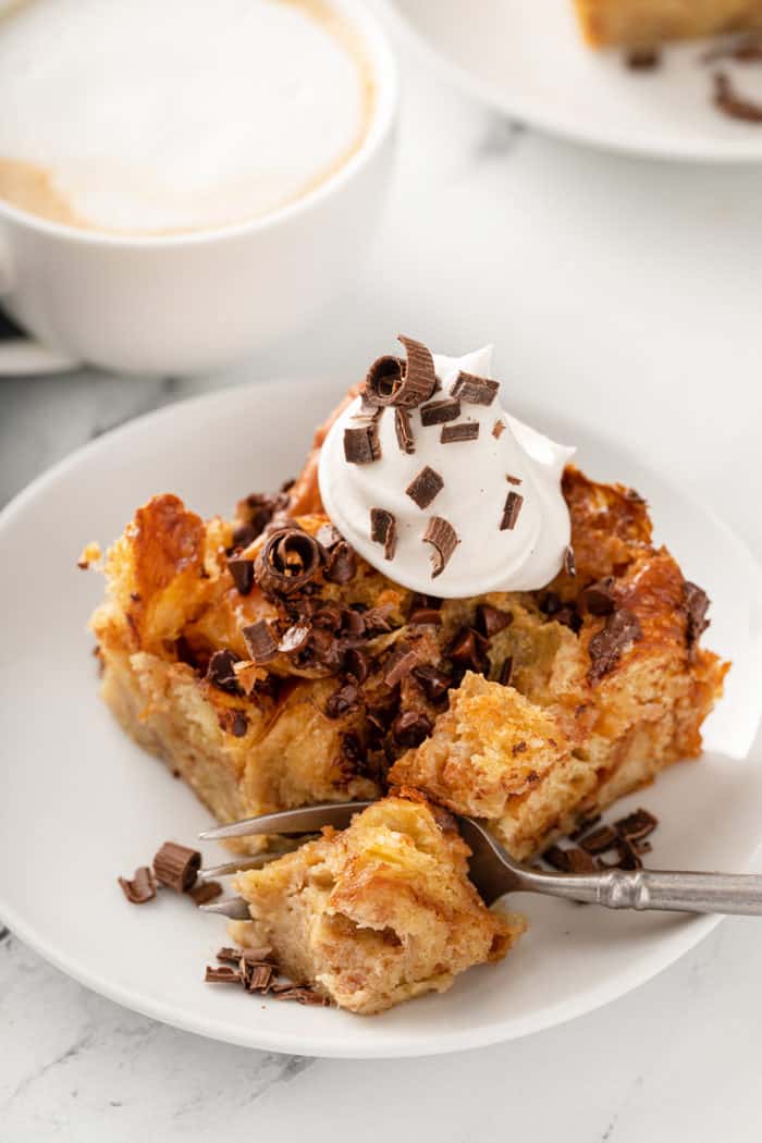 Fork taking a bite from a piece of chocolate chip bread pudding, topped with whipped cream and chocolate shavings