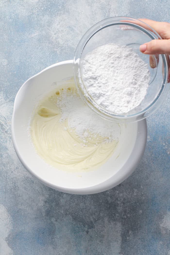 Powdered sugar being added to a white mixing bowl with beaten butter and cream cheese