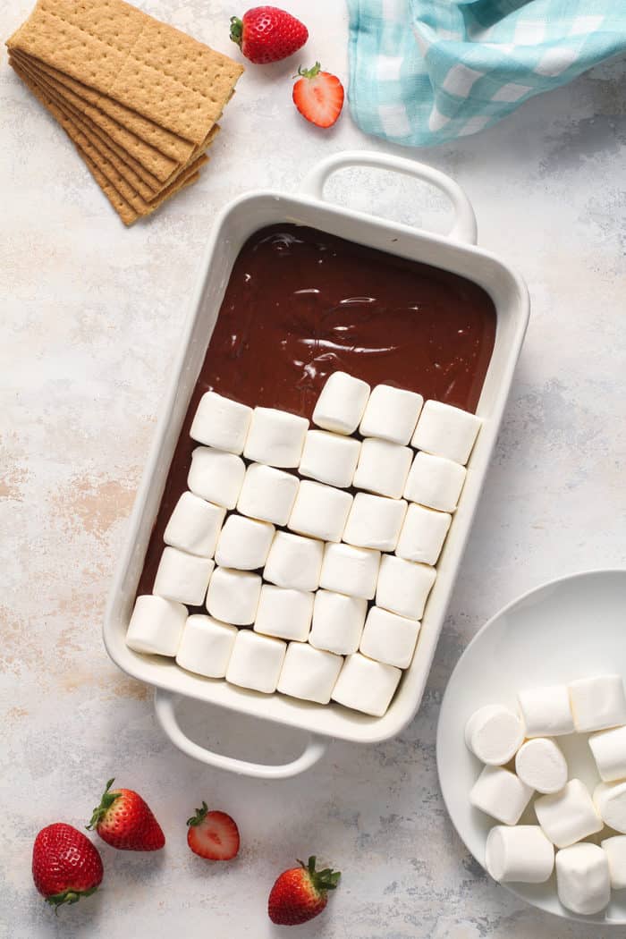 Marshmallows being arranged over the top of chocolate ganache spread across the bottom of a white baking dish