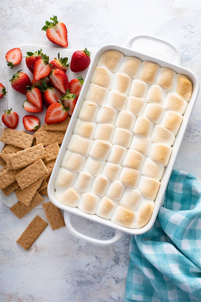 Overhead view of baked s'mores dip in a white baking dish, next to graham crackers and fresh strawberries