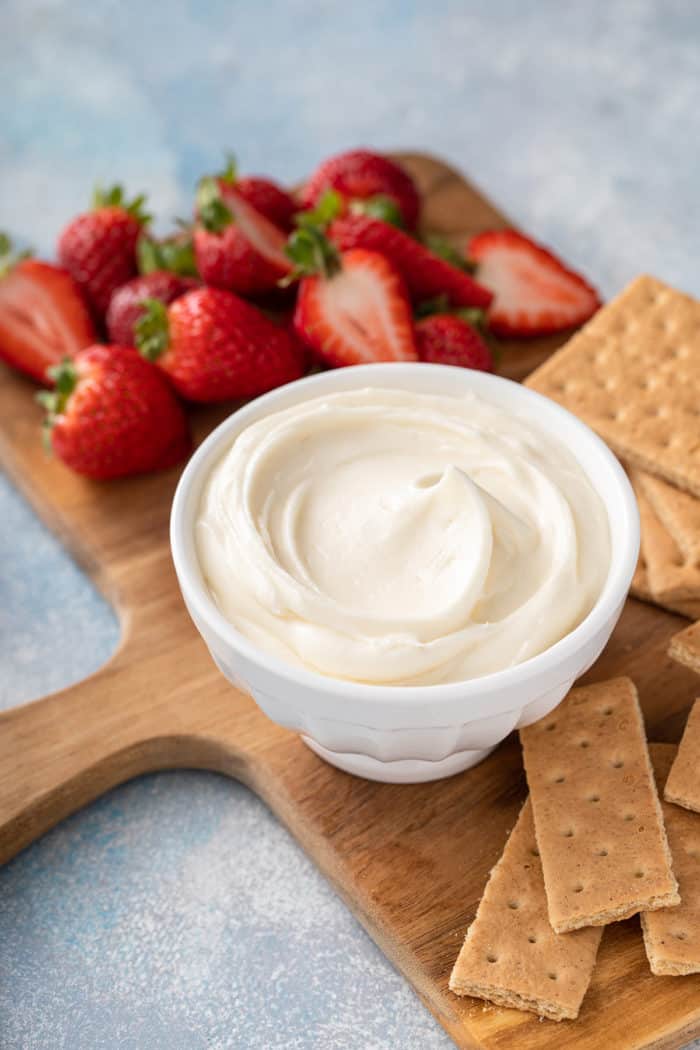 White bowl filled with cream cheese frosting set on a wooden board next to graham crackers and fresh strawberries