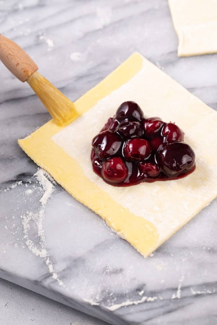 Square of puff pastry with cherry pie filling in the center set on a marble surface. A pastry brush is brushing egg wash along two of the edges of the puff pastry.