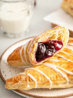 Two cherry turnovers on a white plate. One of the turnovers is cut in half with the cherry filling facing the camera