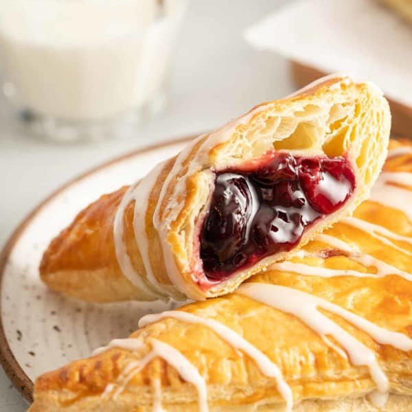 Two cherry turnovers on a white plate. One of the turnovers is cut in half with the cherry filling facing the camera