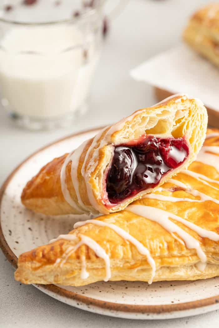 Two cherry turnovers on a white plate. One of the turnovers is cut in half with the cherry filling facing the camera