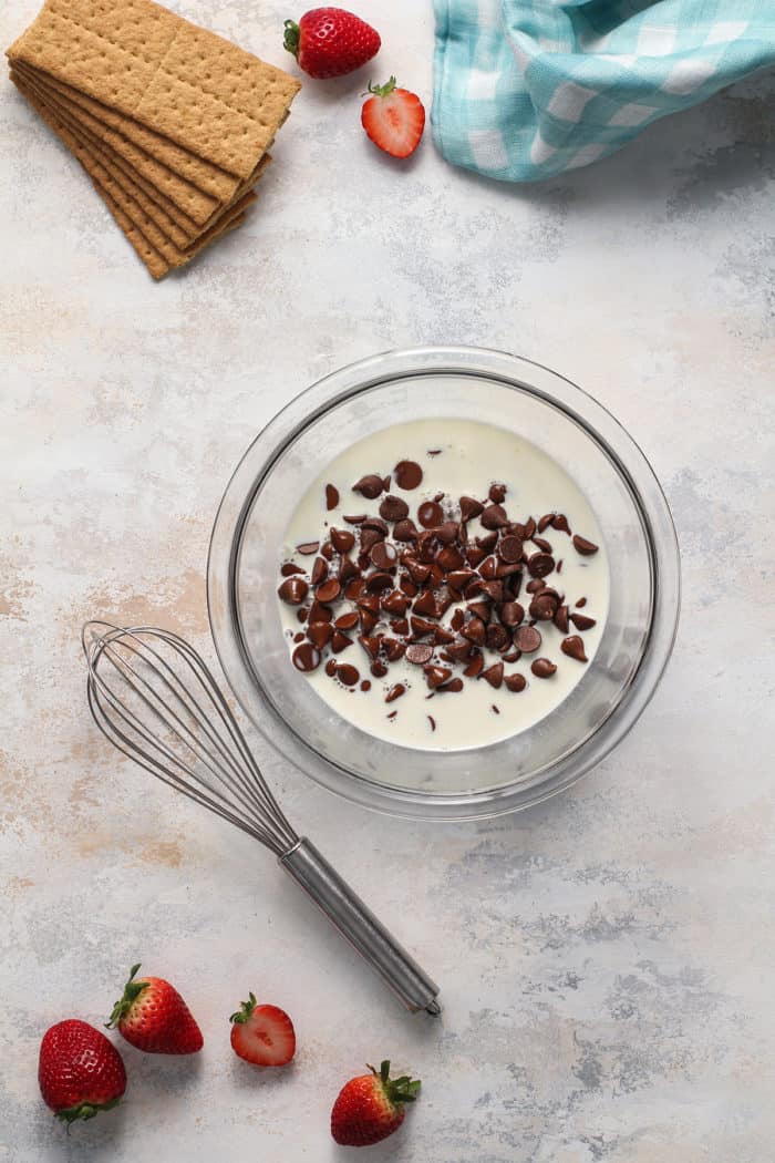 Heavy cream over chocolate chips in a small glass bowl, with a whisk set next to the bowl