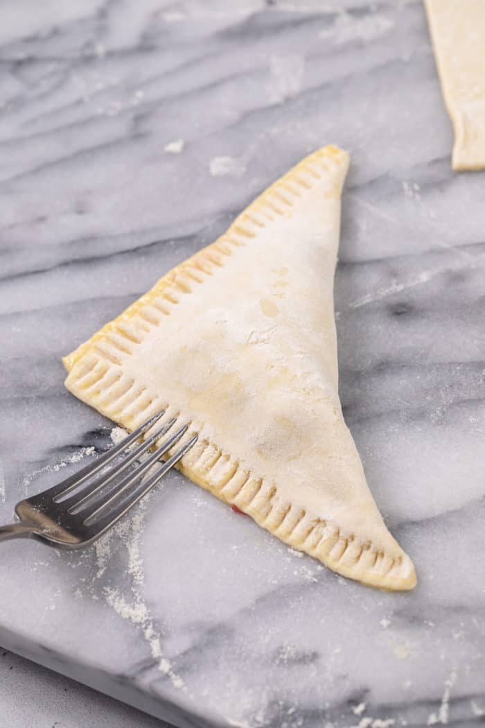 Fork crimping the edges of a cherry turnover set on a marble surface