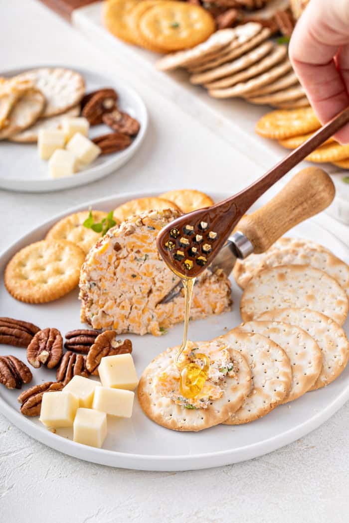 Piece of cheese ball on a plate surrounded by crackers. Honey is being drizzled on top of some of the cheese ball that is spread on a cracker