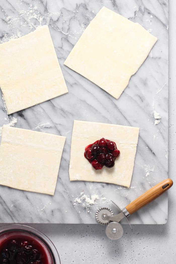 Four squares of puff pastry on a marble surface. One of the squares has cherry pie filling in the center