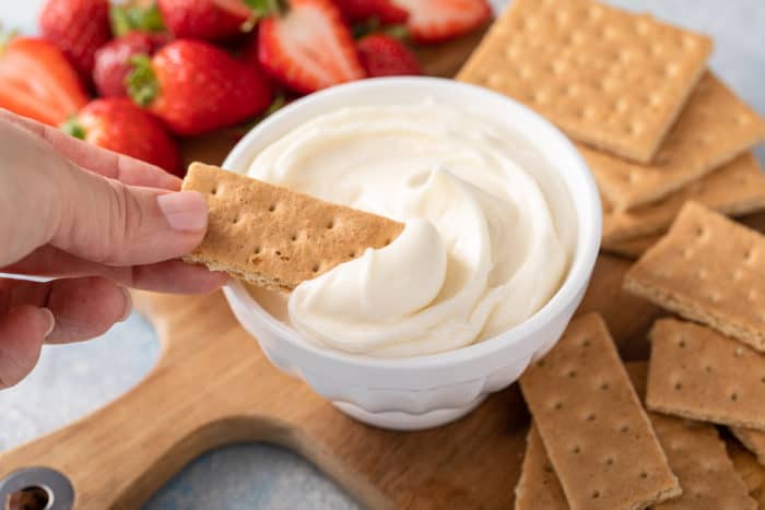 Graham cracker being dipped into a small bowl of cream cheese frosting, surrounded by graham crackers and strawberries