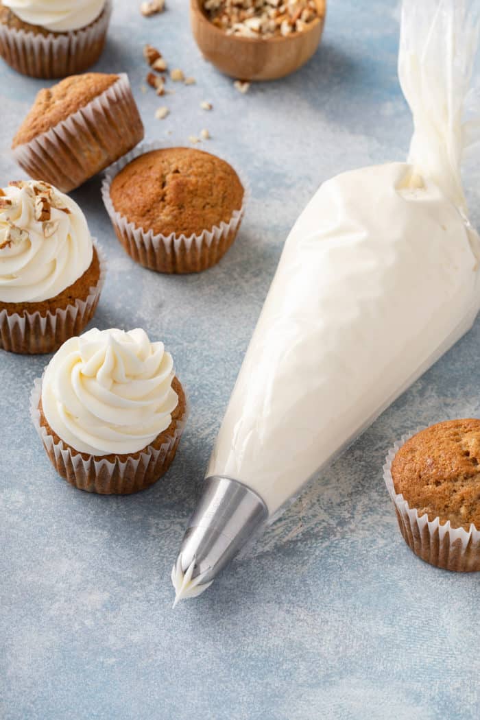 Piping bag filled with cream cheese frosting set on a blue countertop next to cupcakes