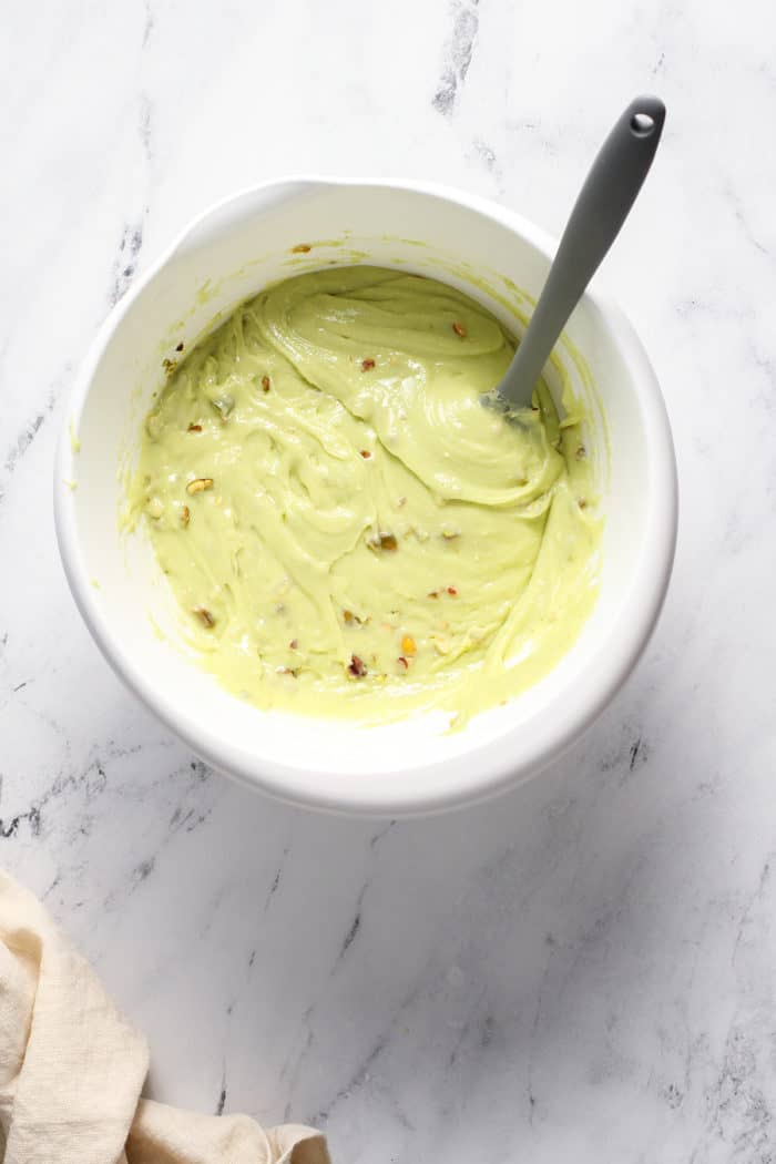 Spatula stirring chopped pistachios into pistachio cake batter in a white mixing bowl
