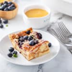 Slice of blueberry bread pudding, dusted with powdered sugar, on a white plate