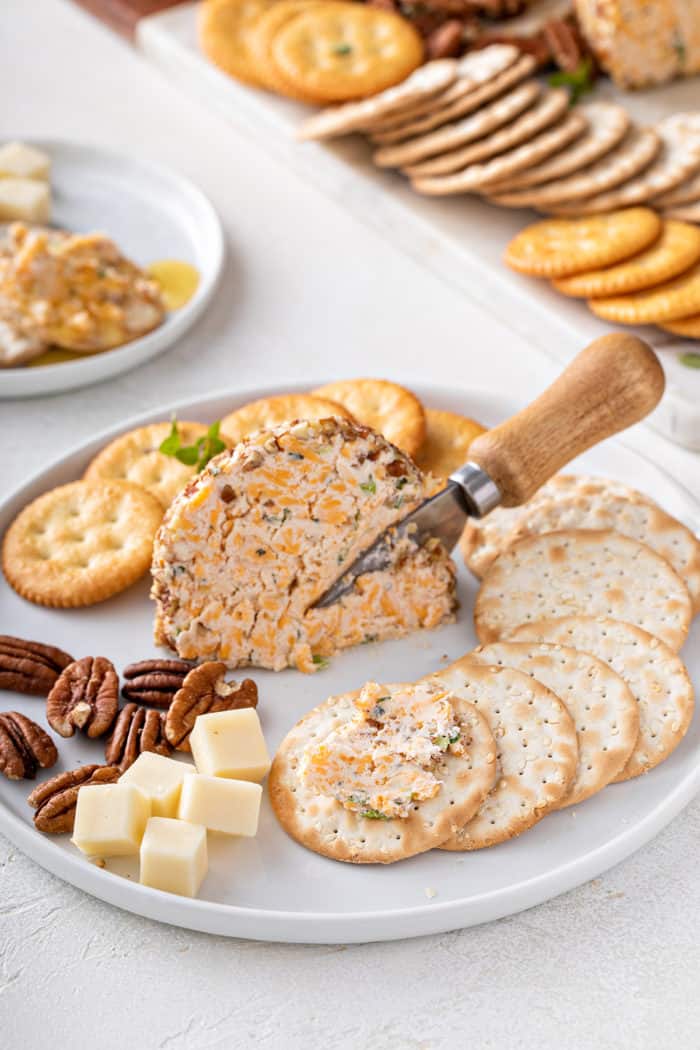Piece of cheese ball on a white plate surrounded by crackers and nuts. Some cheese ball has been spread on one of the crackers