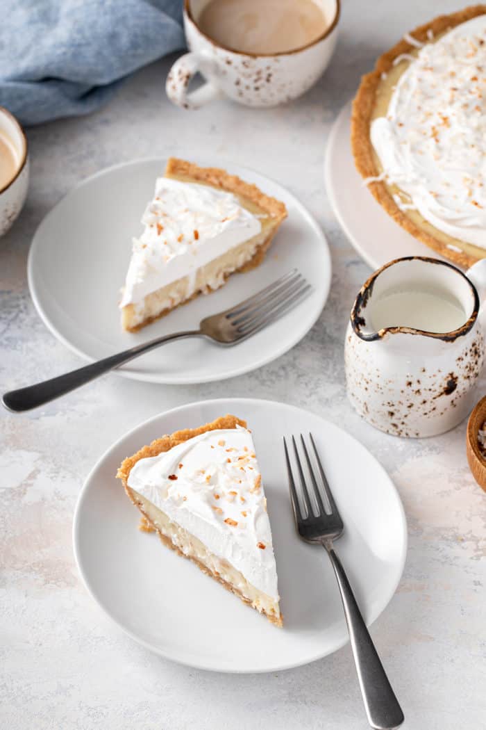 Two white plates with slices of coconut cream pie. A full pie is in the background