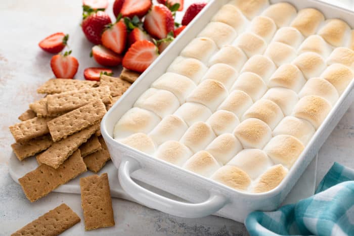 3/4 view of s'mores dip in a white baking dish, set next to graham crackers and fresh strawberries