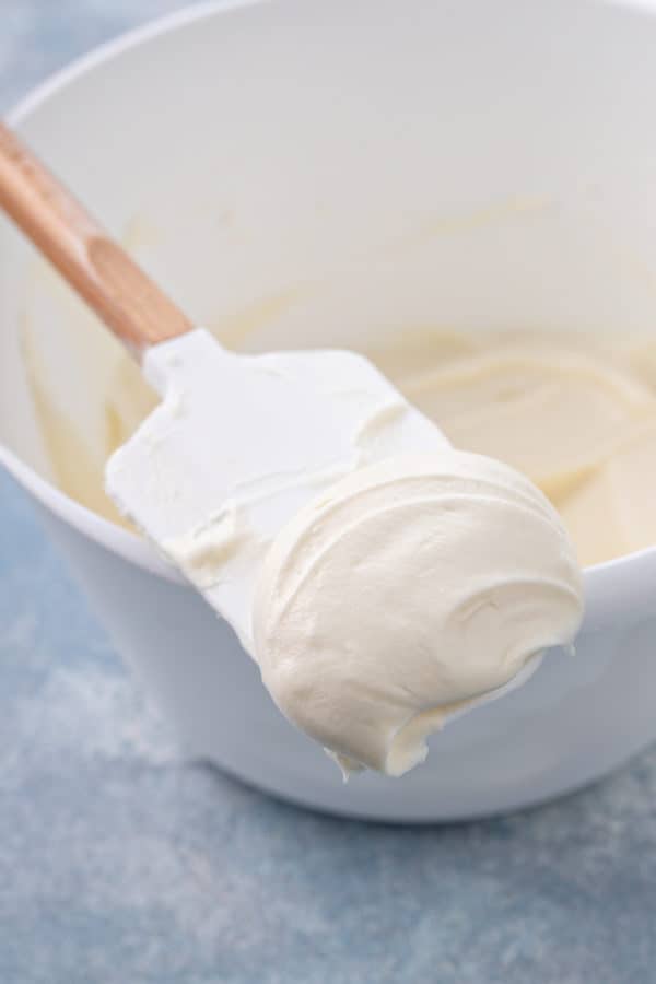 Spatula with cream cheese frosting on it set on the side of a white mixing bowl