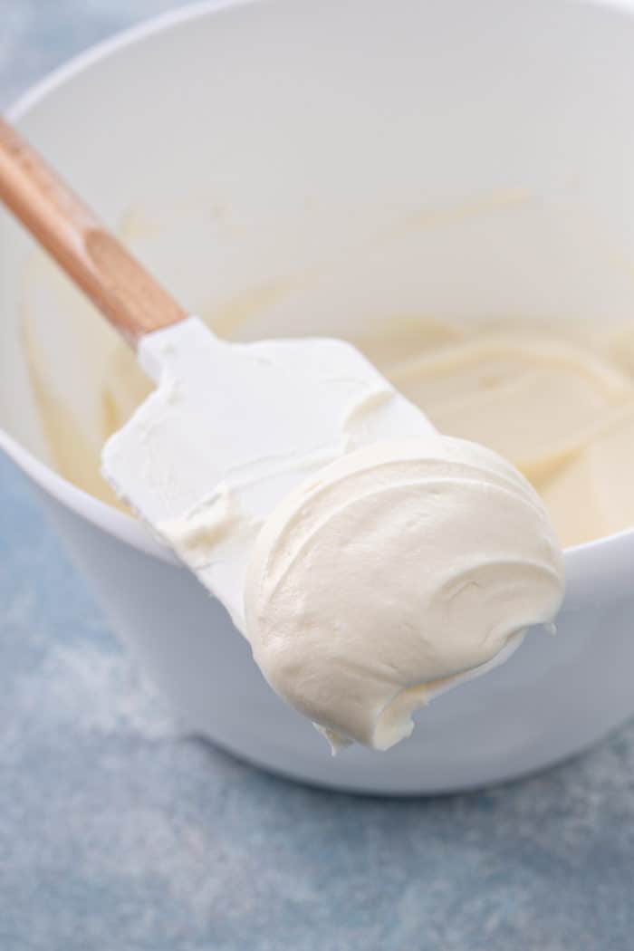 Spatula with cream cheese frosting on it set on the side of a white mixing bowl