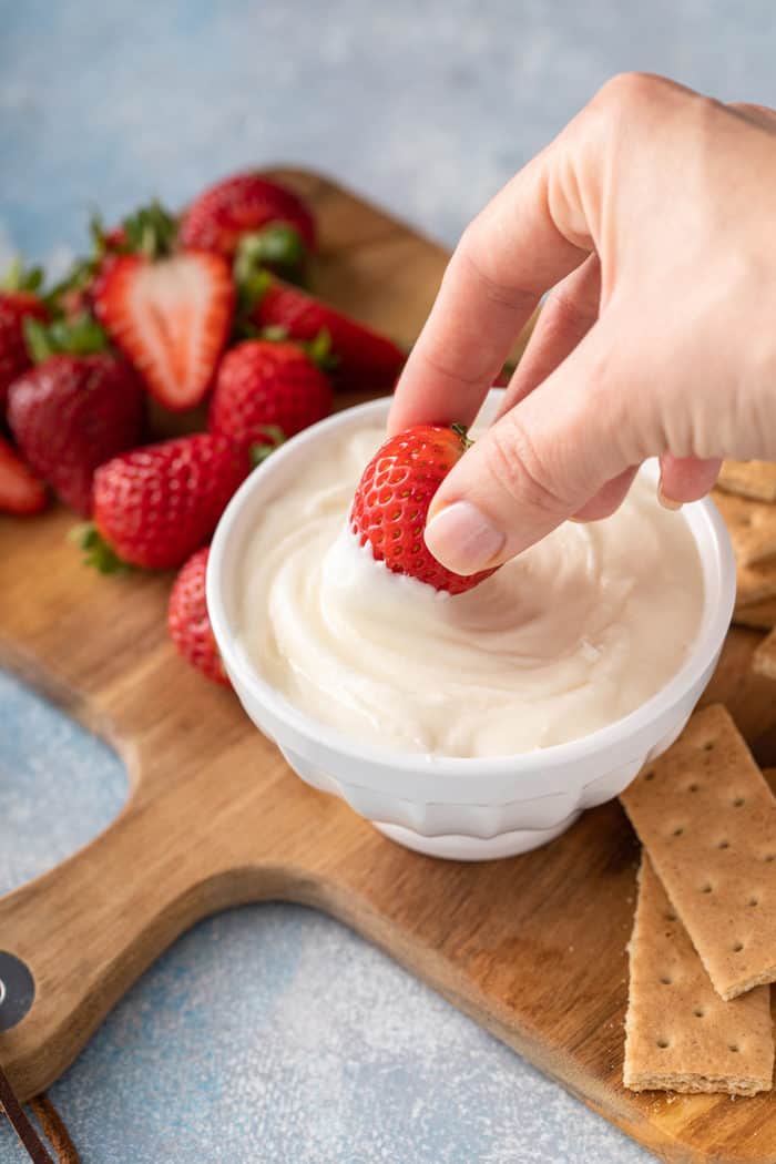 Hand dipping a fresh strawberry into a bowl of cream cheese frosting