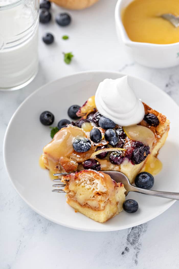 Fork taking a bite from a slice of blueberry bread pudding that is topped with lemon curd and whipped cream