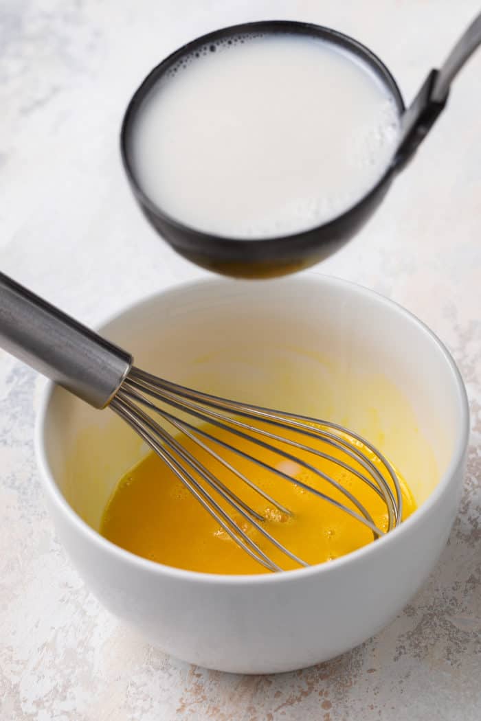 Hot milk being added to whisked egg yolks in a small white bowl