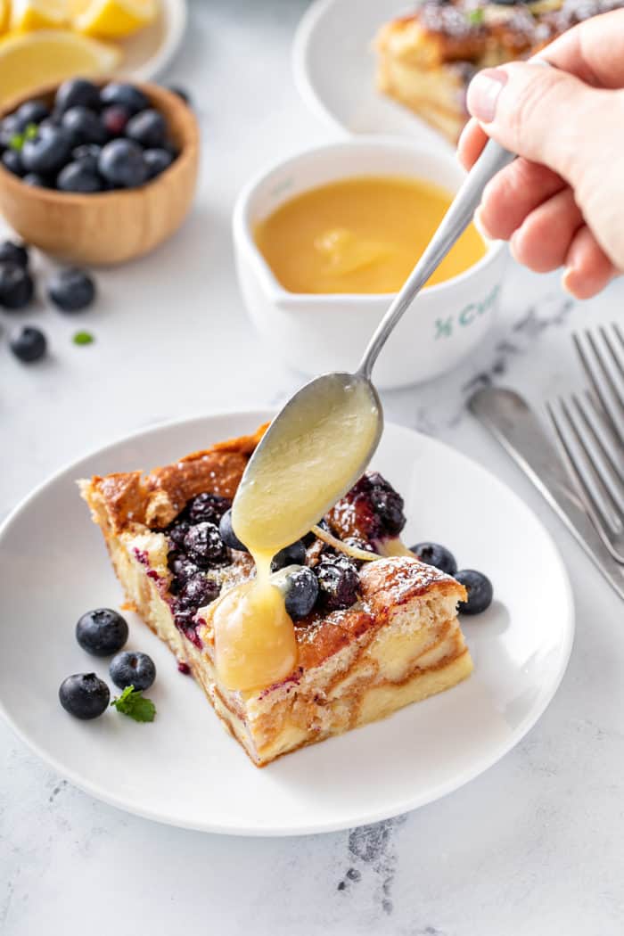 Spoon dolloping lemon curd over the top of a slice of blueberry bread pudding on a white plate