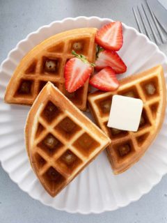 Three bisquick waffles on a white plate with syrup, butter and fresh strawberries