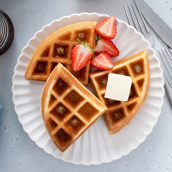 Three bisquick waffles on a white plate with syrup, butter and fresh strawberries