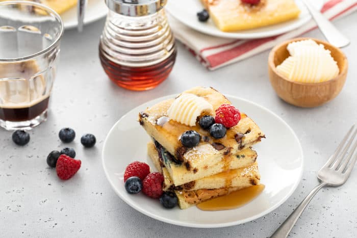 Stack of sheet pan pancakes on a white plate, topped with butter, berries, and syrup