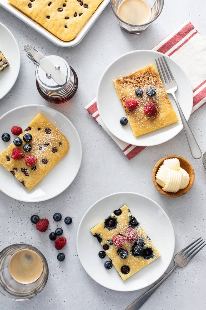 Overhead view of three sheet pan pancakes on white plates, topped with peanut butter, blueberries, and chocolate chips