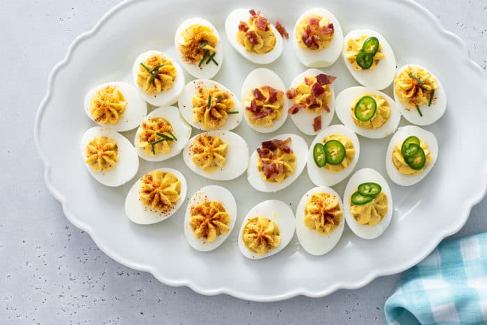Assorted deviled eggs on a white platter, topped with chives, bacon, and jalapeños
