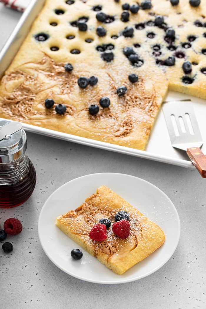 Slice of sheet pan pancake on a white plate with the pan of pancakes in the background