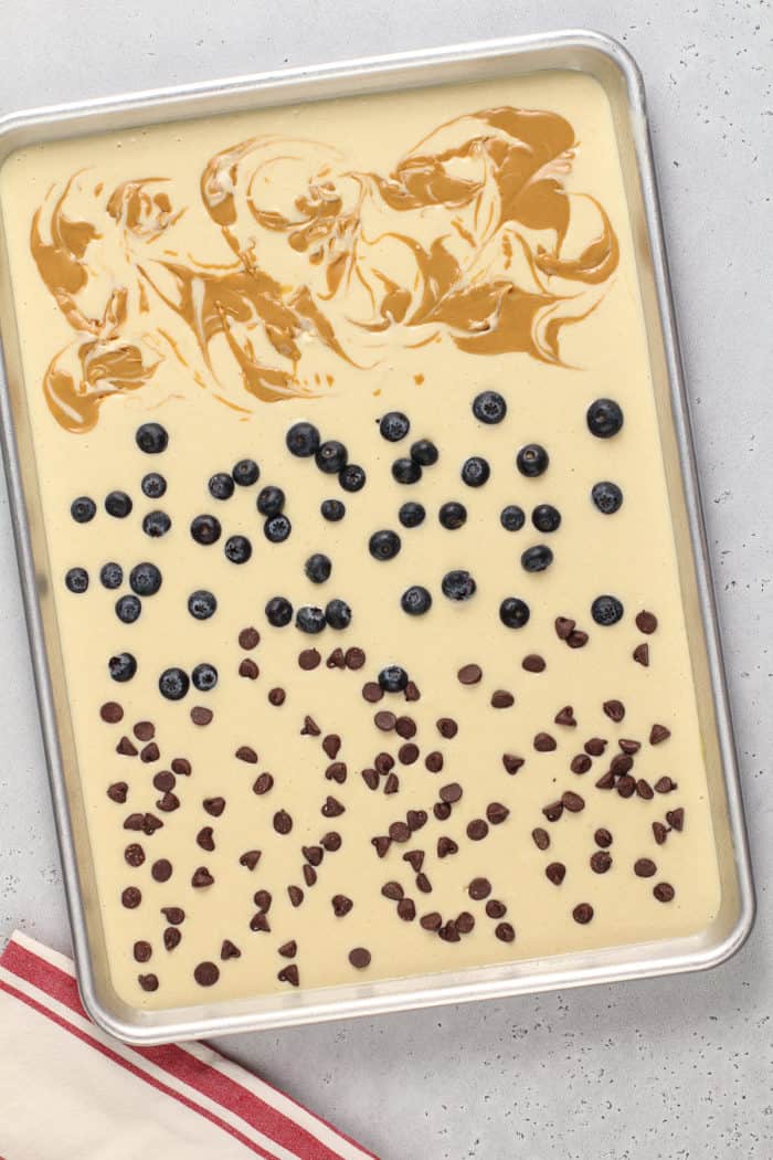 Pancake batter in a sheet pan, topped with peanut butter, blueberries and chocolate chips