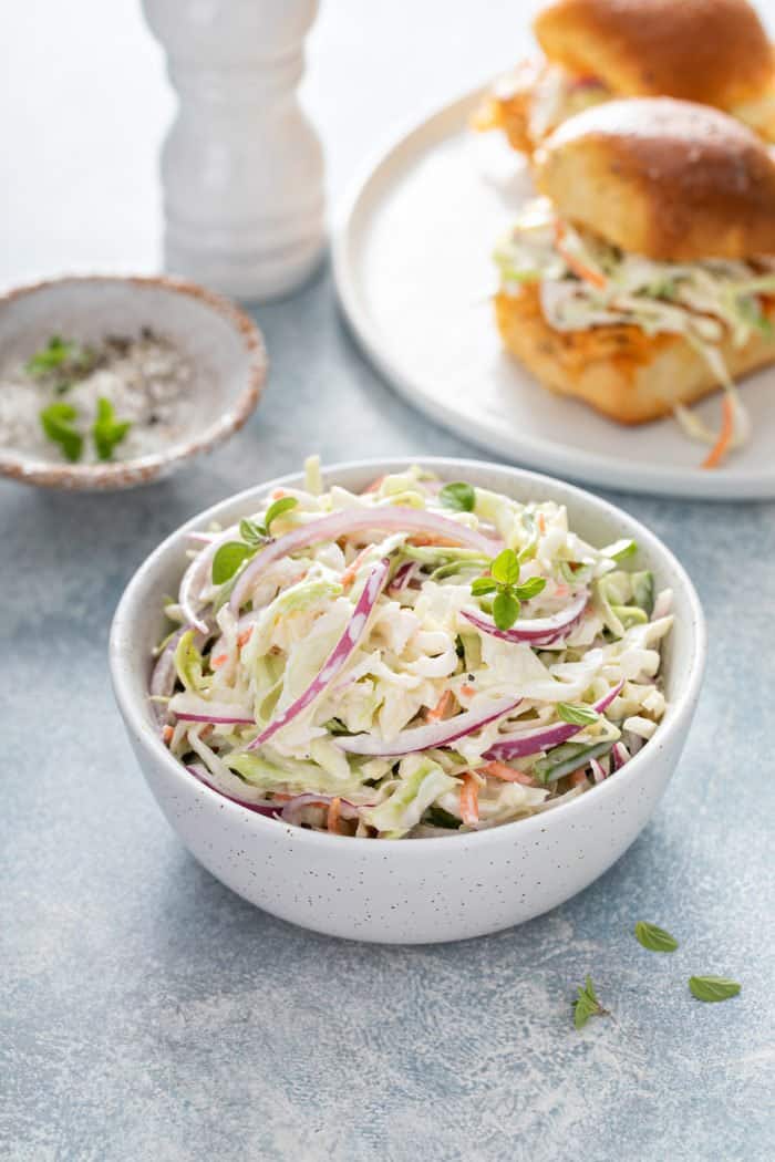 Small white bowl of easy coleslaw with a plate of sliders in the background