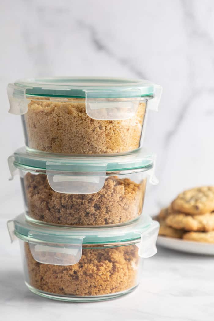 Three airtight containers filled with brown sugar stacked on a marble countertop with a plate of cookies in the background
