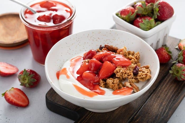 White bowl filled with yogurt, granola, and homemade strawberry sauce, set on a wooden cutting board
