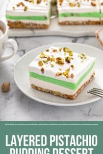 Slice of pistachio pudding dessert on a white plate, with the rest of the layered dessert in the background.