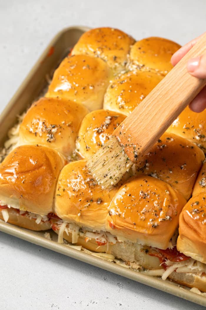 Herbed butter being brushed over the tops of unbaked pizza sliders.