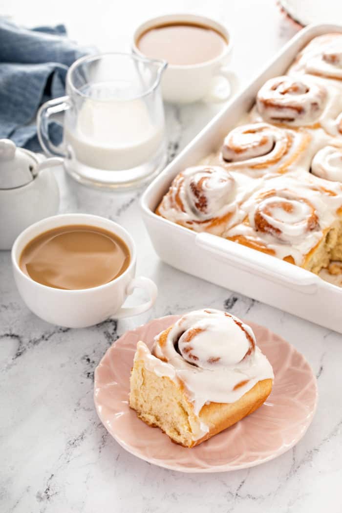 Plated overnight cinnamon roll set in front of a cup of coffee and the pan of cinnamon rolls
