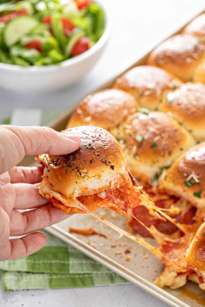 Hand pulling a pepperoni pizza slider off a baking sheet.