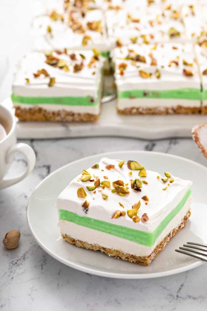 Slice of pistachio pudding dessert on a white plate, with the rest of the layered dessert in the background.