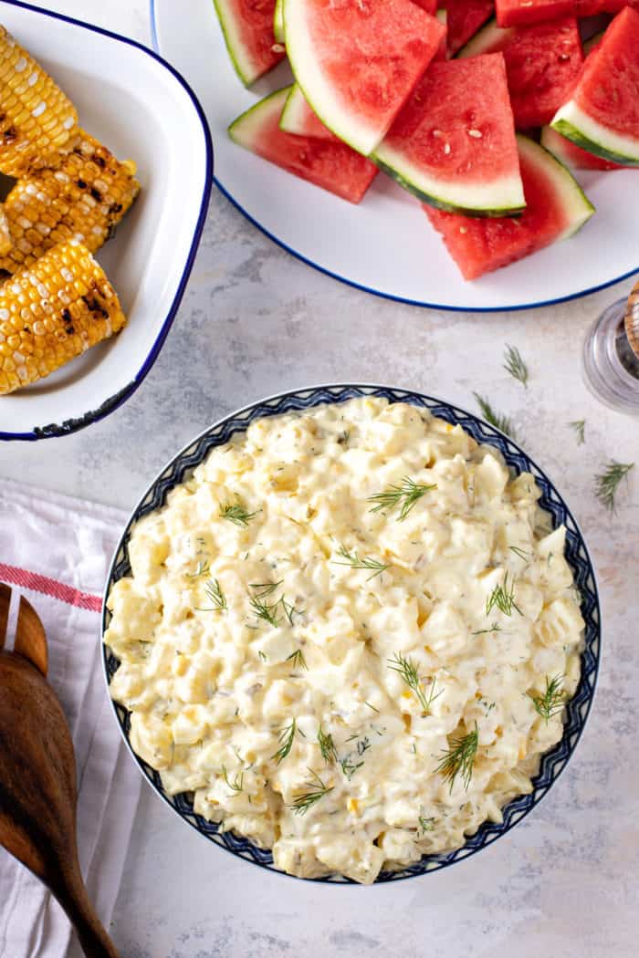 Overhead view of a serving bowl filled with easy potato salad next to platters of fresh watermelon and grilled corn.