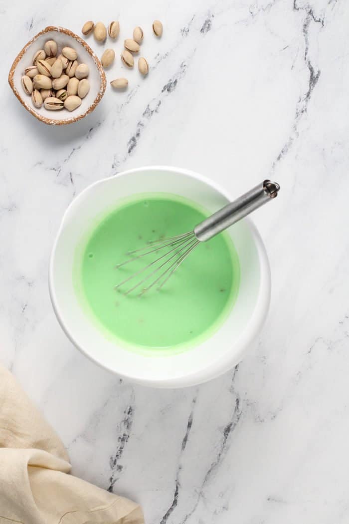 Pistachio pudding being whisked together in a white mixing bowl on a marble countertop.