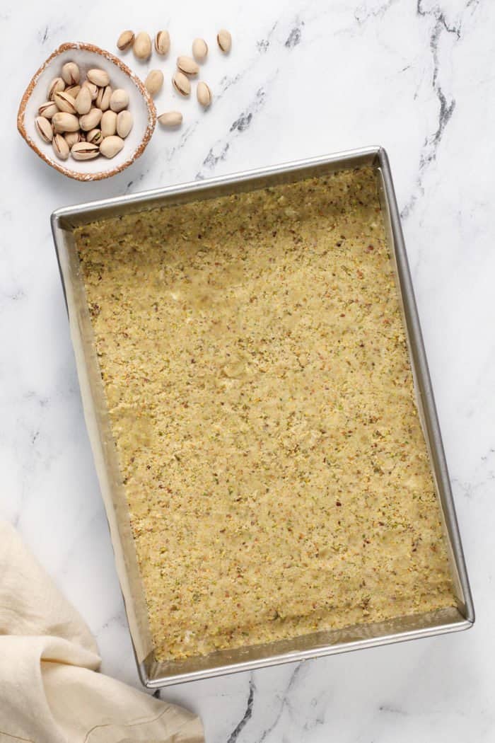 Crust for pistachio pudding dessert in the bottom of a metal pan, set on a marble countertop.