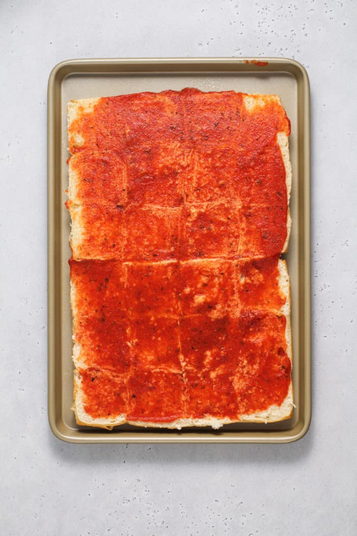 Pizza sauce spread on the bottoms of hawaiian roll slider buns set on a rimmed baking sheet.