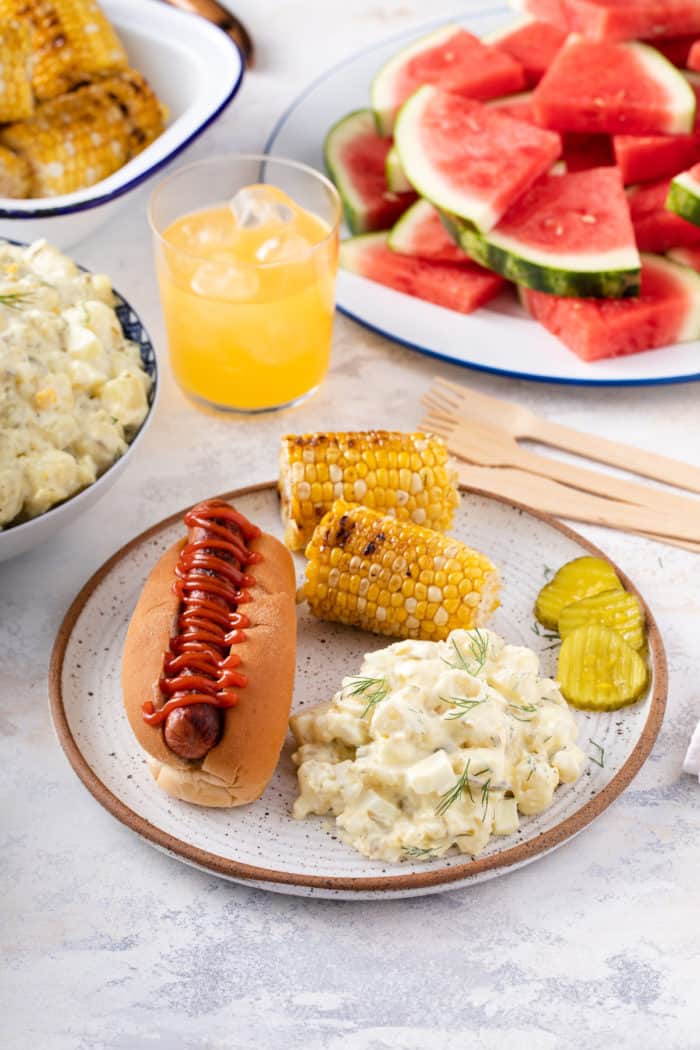 Table filled with summer cookout food, with a plate of potato salad, a hot dog, and corn in the foreground.