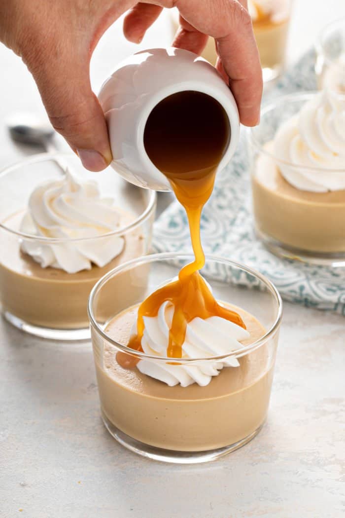 Hand pouring caramel sauce over a bowl of butterscotch pudding topped with whipped cream.